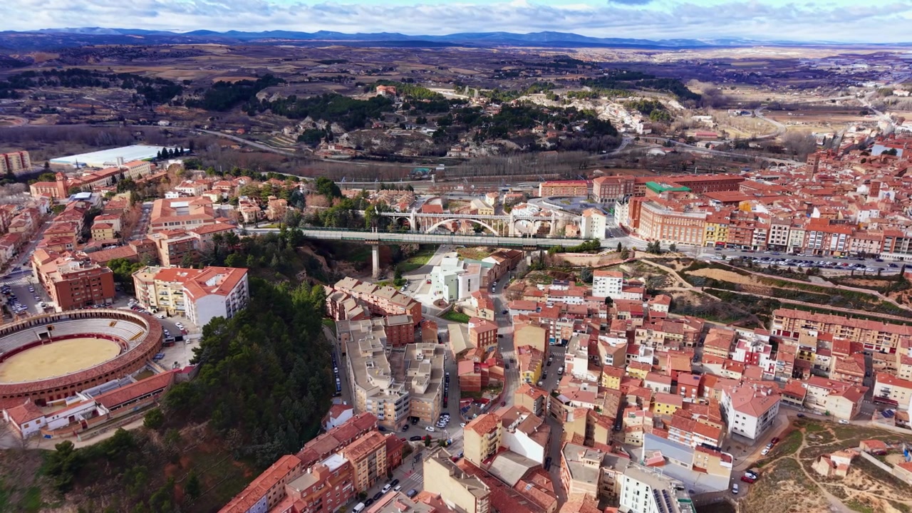 Vista panoramica di Teruel, Spagna, che mostra tetti in terracotta, un'arena per la corrida, un ponte storico e il paesaggio della valle circostante sotto un cielo nuvoloso.