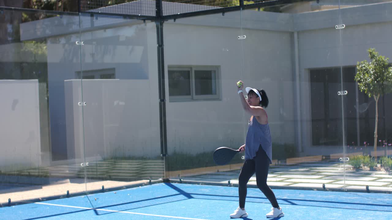 Mature African American woman bouncing ball, serving and swinging paddle on blue court, copy space