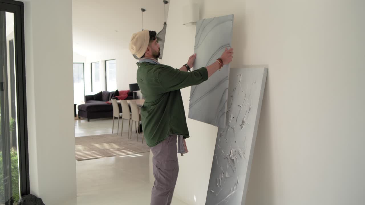 Uomo indiano che entra nel corridoio indossando un berretto di lana, rimuovendo un quadro appeso e riappendendo una nuova tela a un gancio a parete.
