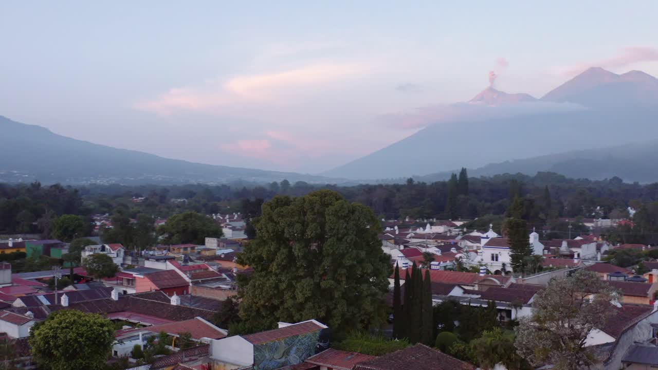 Bredt, dynamisk droneoptagelse kredser om et massivt træ, der tårner sig op over Antigua, Guatemala, med tre synlige vulkaner i baggrunden: Agua, Acatenango og Fuego, hvoraf den sidste er i synligt udbrud