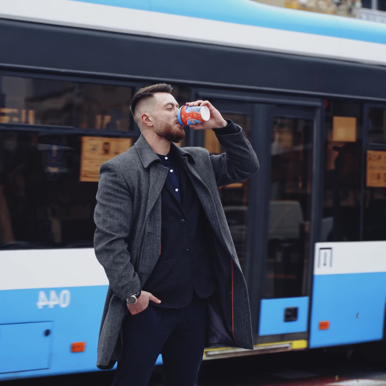 Handsome man in the city. Young businessman waiting for someone on the urban traffic background. Man in coat holds a plastic glass with coffee and looks on watch.