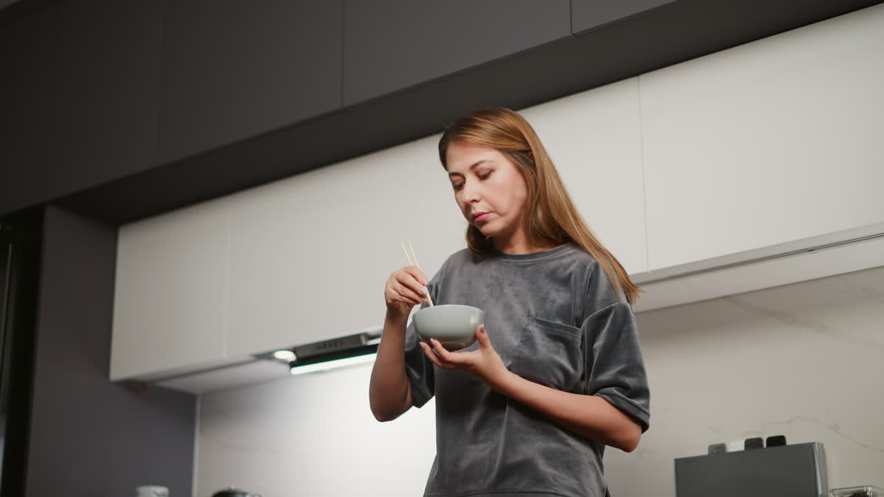 Young woman with long flowing hair and painted lips stands in modern kitchen eating lunch from bowl using chopsticks, bright under cabinet light illuminating