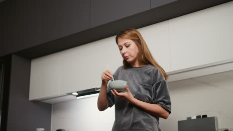 young woman with long flowing hair and painted lips stands in modern kitchen eating lunch from bowl using chopsticks, bright under cabinet light illuminating