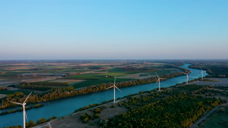 Tiro-De-Seguimiento-Aéreo-Desde-Arriba-De-Cinco-Turbinas-Eólicas-Cerca-De-Un-Río-En-El-Sur-De-Francia-Al-Atardecer