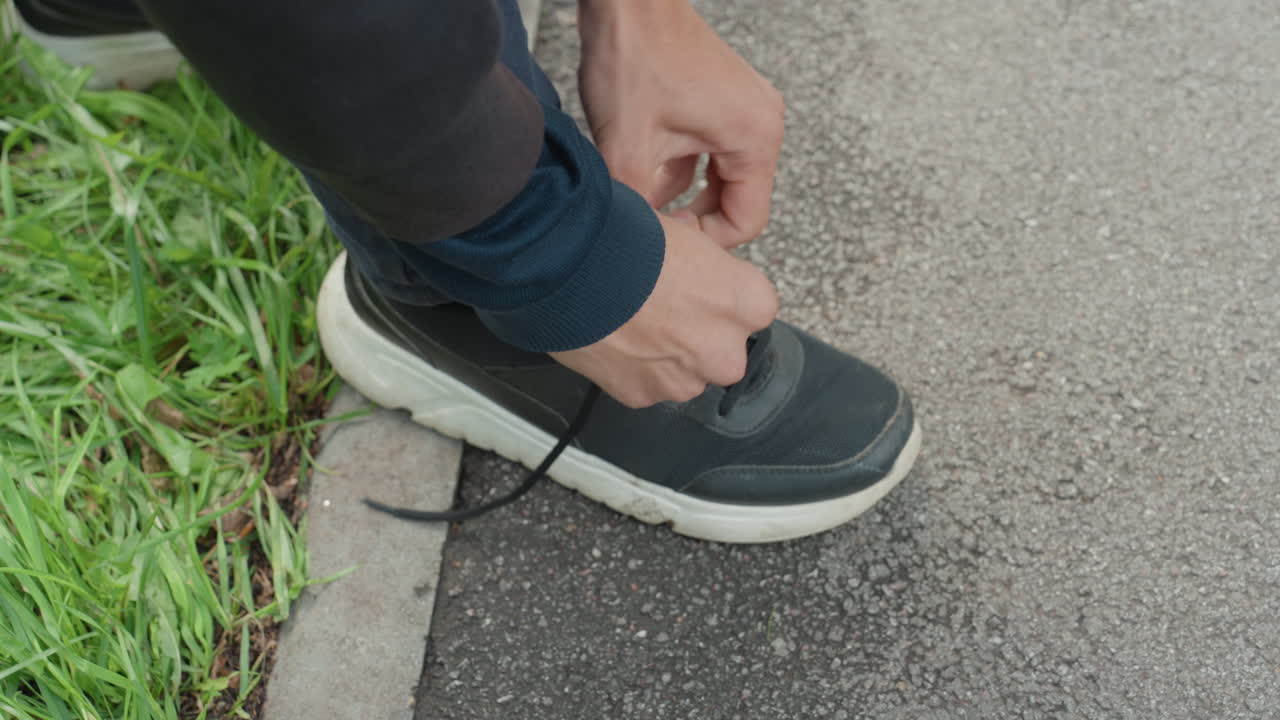 primo piano di una scarpa sul marciapiede mentre allaccia i lacci vicino all'erba, mani che sistemano i lacci con gesto concentrato, suola testurizzata sul pavimento bagnato, jeans casual e sneaker scure, calma preparazione mattutina e pausa consapevole
