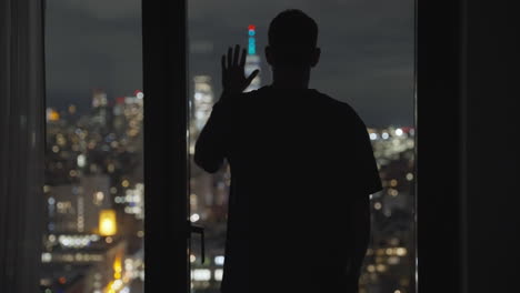 Lonely-man-standing-with-back-to-camera-in-silhouette-against-metropolitan-cityscape-at-night-watching-the-city-in-sad-emotional-look