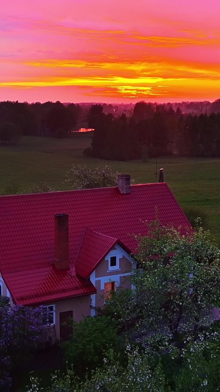 Flyfoto over et landlig hus og en dam ved solnedgang, omgitt av grønne trær og jorder.