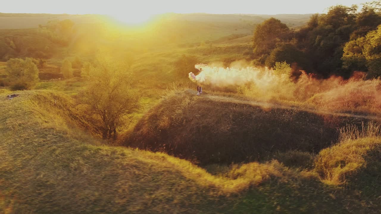 Couple With Smoke Bombs. Aerial shot of a happy couple walking through the field with smoke bombs on the background of sunset