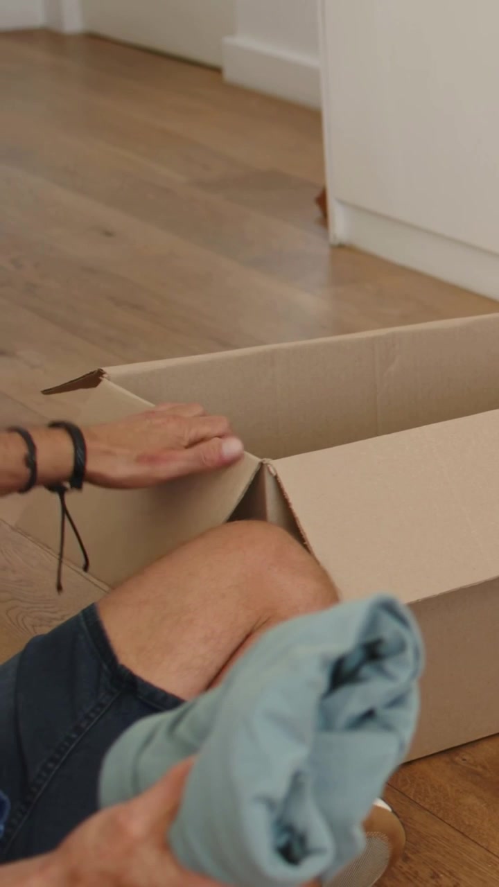Video vertical: Hombre con camisa azul entrando y metiendo un cojín en una caja en casa para empacar, con espacio para texto.