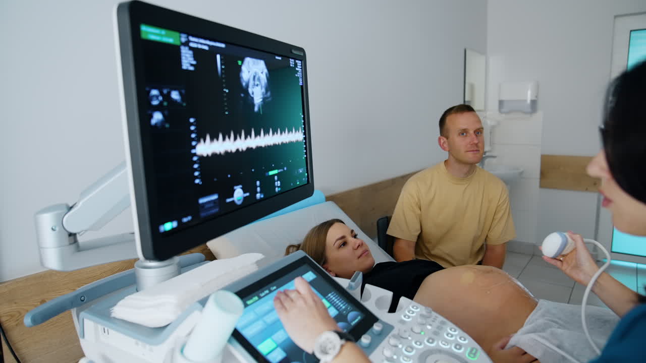 Brunette female doctor sits looking at the screen of ultrasonic machine. Doctor is commenting on the baby development to couple at check up.