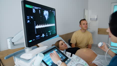 brunette female doctor sits looking at the screen of ultrasonic machine. doctor is commenting on the baby development to couple at check up.