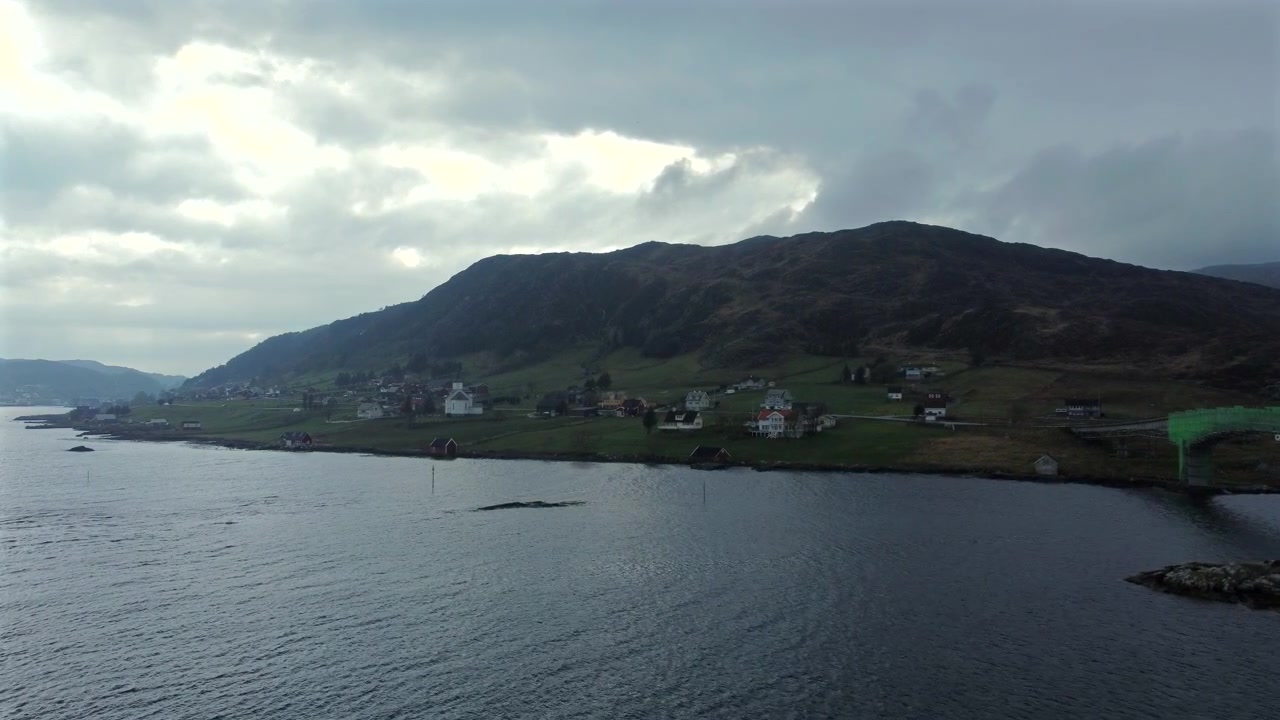 Fremadrettet luftfoto over en sø mod en bakkeskråning med spredte huse og grønne marker i Norge.