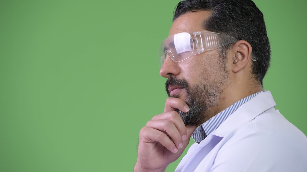 Vista di profilo di un medico persiano barbuto bello che indossa occhiali protettivi e pensa