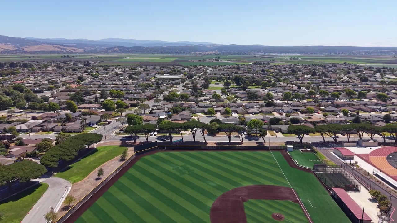 Aerial footage of Hartnell College in Salinas, California, showing sports fields, neat suburban homes, and distant farmland on a clear, sunny day. Vibrant sky, smooth drone movement.