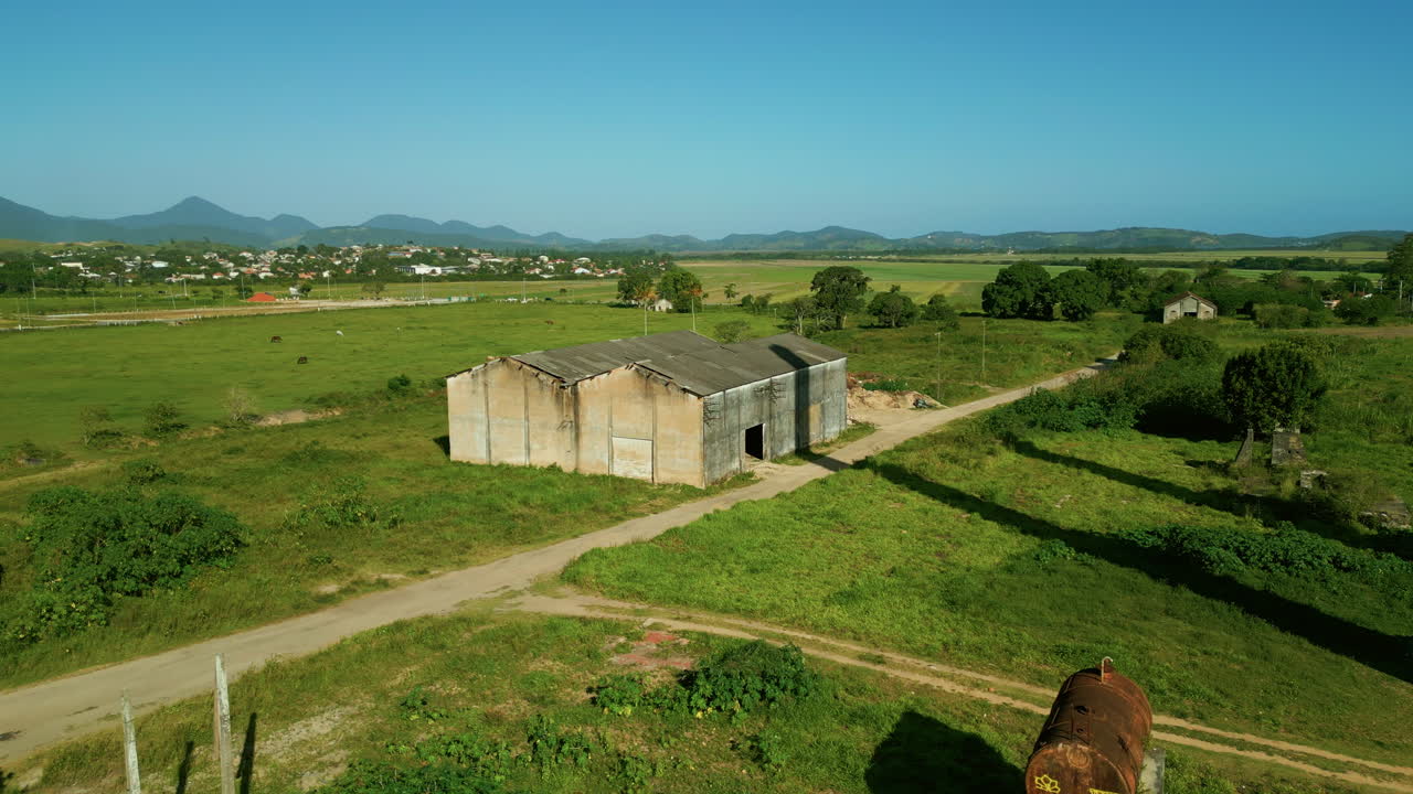 Un drone che vola a bassa quota si muove in avanti sopra una casa abbandonata verso il zuccherificio e distilleria di etanolo di Santa Luiza a Saquarema, in Brasile. Rovine industriali, campi verdi e ciminiere gemelle dominano il paesaggio rurale