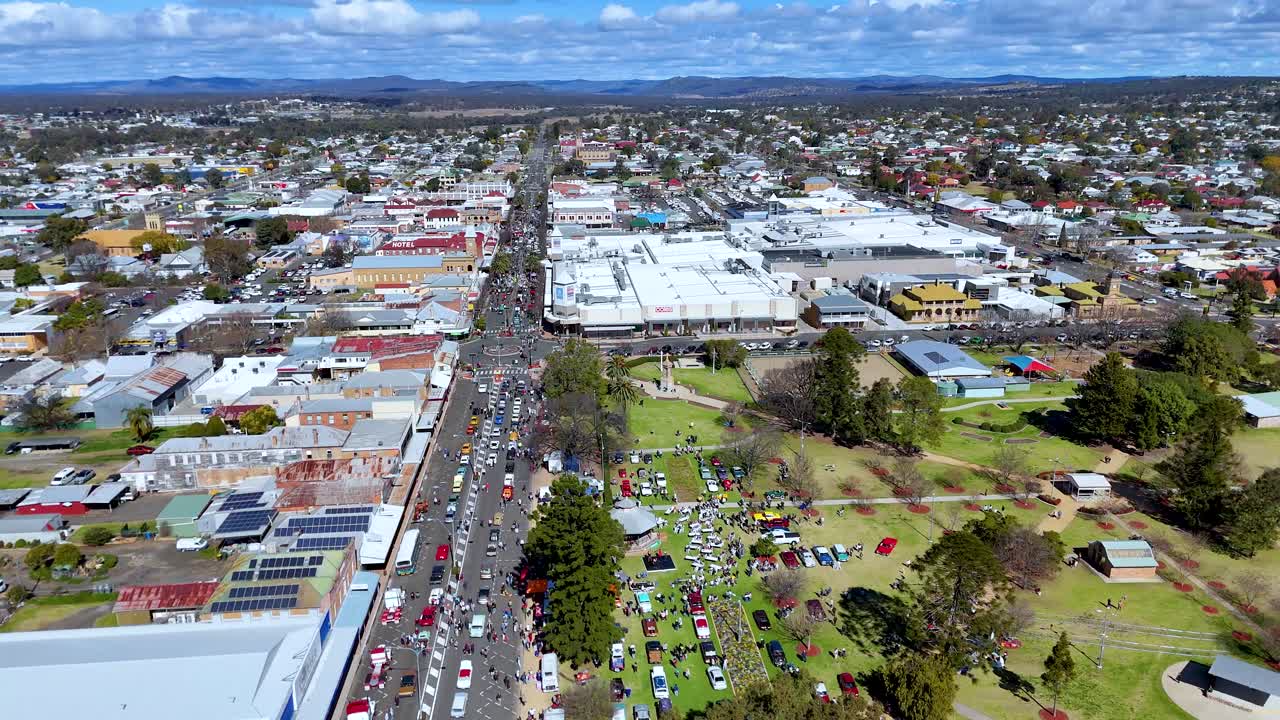 Drone footage glides above a vibrant town street lined with festival stalls, crowds, and parked cars on a sunny day, capturing community atmosphere