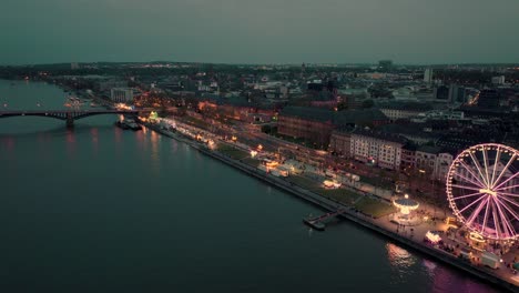 Toma-Aérea-De-Un-Dron-De-Maguncia-Retirándose-De-Un-Festival-Del-Vino-Que-Muestra-Toda-La-Ciudad-En-Un-Amplio-Plano-Sobre-El-Río-Rin-En-Una-Puesta-De-Sol-De-Verano-Con-Muchas-Luces-De-La-Ciudad