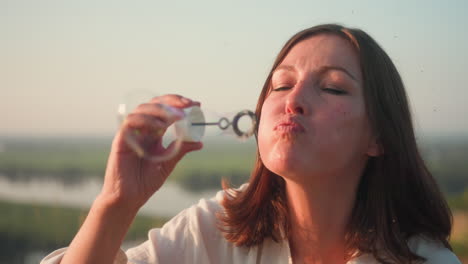 young woman blowing soapy bubbles into sky while seated on grassy hill overlooking river valley at sunset, warm light illuminating hair and bubbles floating in peaceful natural setting