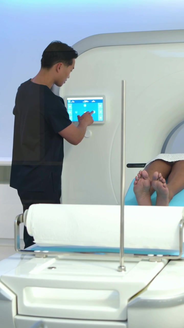 Video verticale: Regolazione della testa del paziente, un tecnico in camice prepara il pannello dello scanner, paziente in camice.