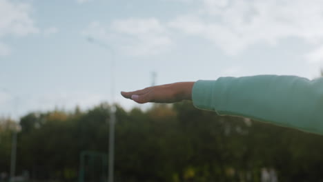 Elegant woman performing hand stretches during outdoor fitness session on green grassy field under bright sunlight with serene trees in background creating peaceful and motivating environment