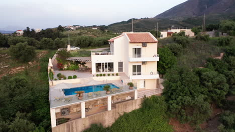 Vuelo-Aéreo-En-El-Sentido-De-Las-Agujas-Del-Reloj-Alrededor-De-Una-Villa-Griega-De-Lujo-Con-Piscina-Con-Montañas-En-El-Fondo-Y-Follaje-Verde-En-Primer-Plano---Coche-En-Marcha