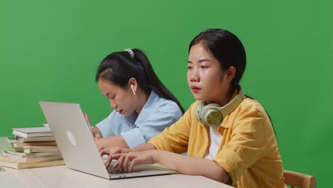 close up of unsatisfied asian woman student sitting with her friend and typing on a laptop on the table in the green screen background classroom