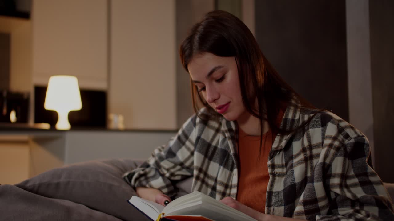 A happy brunette girl in a plaid shirt and an orange T-shirt reads a book and flips through its pages while relaxing on a gray sofa in a modern apartment in the evening