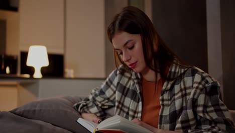 a happy brunette girl in a plaid shirt and an orange t-shirt reads a book and flips through its pages while relaxing on a gray sofa in a modern apartment in the evening