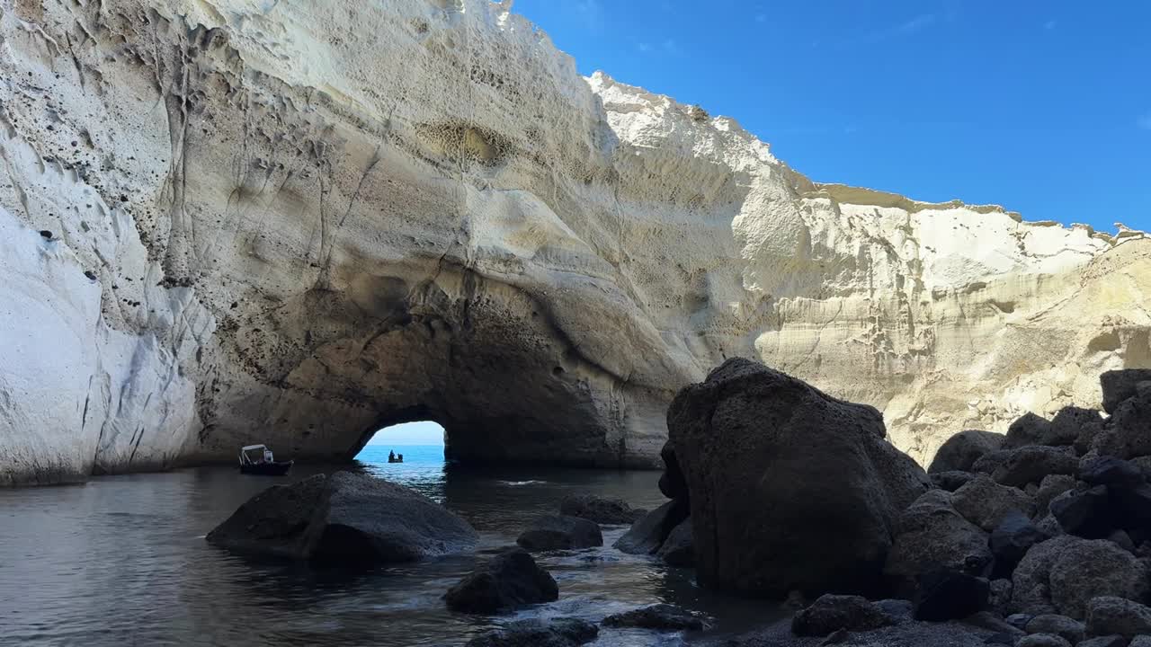 Boat entering the entrance to Sikia Cave in Milos glows under the bright sun on a clear day.