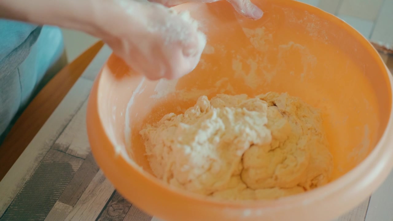 Close up shot of teenage hands kneading fresh sticky dough inside an orange bowl. Traditional bread making process, working with texture, culinary skills, cozy home kitchen lifestyle, 4k.