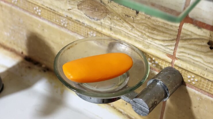 Close-up of an orange bar of soap resting on a clear glass soap dish mounted to a tiled bathroom wall beside a sink, showing everyday hygiene, cleaning routine, and indoor washroom details