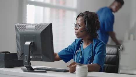 médica general negra afroamericana con bata blanca sentada en el escritorio de la oficina del médico y desplazando el ratón de la computadora mientras lee el historial médico del paciente