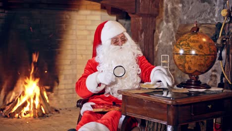 santa claus is sitting in specially decorated room at the table, holding a magnifying glass in hands. feast fireplace in the background. christmas spirit concept close up video. christmas eve. new year is coming. santa claus is looking at the globe with a magnifying glass.