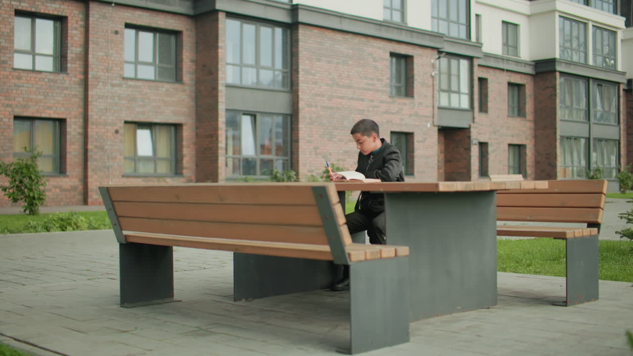 Boy seated at outdoor bench studying attentively with open book flipping in wind, pen in hand, brick residential building in background