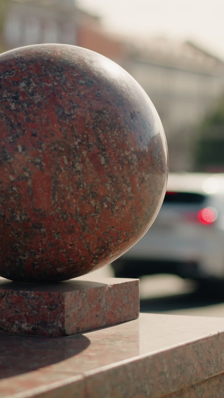 Esfera de granito sobre la acera de la ciudad en primer plano, piedra roja erosionada posada sobre una base cuadrada, tráfico difuminado y edificios históricos al fondo, luz cálida de la hora dorada que crea reflejos suaves y largos