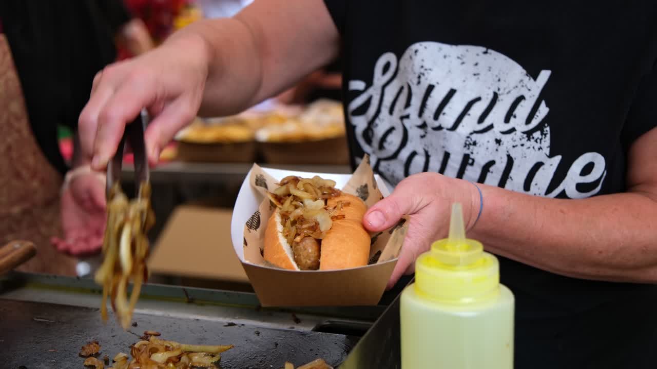 Close up of street food vendor preparing sausage in bun with grilled onions and peppers at market stall. Hands use tongs to serve hotdog into tray, highlighting fresh and tasty festival food culture