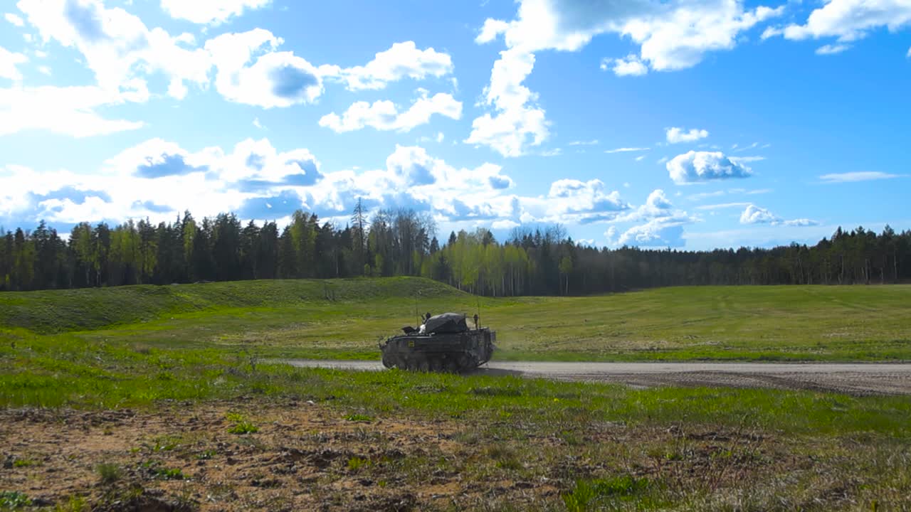 British warrior FV510 armoured peronnel carrier tank driving from a grassy green field to a gravel road in slow motion and creating a dust field, while a soldier is on top of the vehicle. Sunny day.