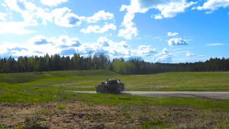 british warrior fv510 armoured peronnel carrier tank driving from a grassy green field to a gravel road in slow motion and creating a dust field, while a soldier is on top of the vehicle. sunny day.