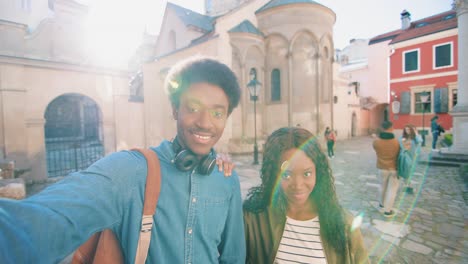 mujer afroamericana y hombre sosteniendo un teléfono inteligente y saludando a la cámara mientras sonríe en la calle