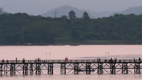 Mon-Bridge-and-a-longboat-followed-speeding-behind-the-structure-while-people-are-walking-on-the-bridge,-silhouetting-as-it-was-getting-dark,-in-slow-motion