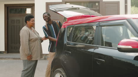 young man and his grandma standing outside next to car with opened trunk talking together before boy moving home