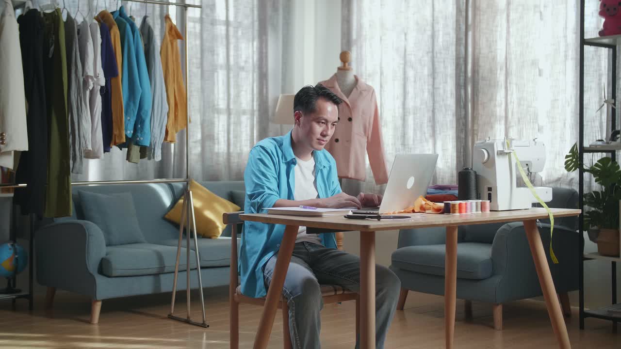 아시아 남성 디자이너가 스튜디오에서 옷을 디자인하는 동안 노트북에서 일하고 있습니다.