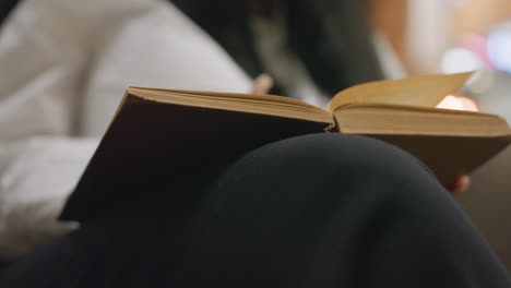 close up woman with long black hair seated flipping book pages on her knee under soft warm bokeh lighting background in cozy lounge setting suggesting peaceful evening reading time
