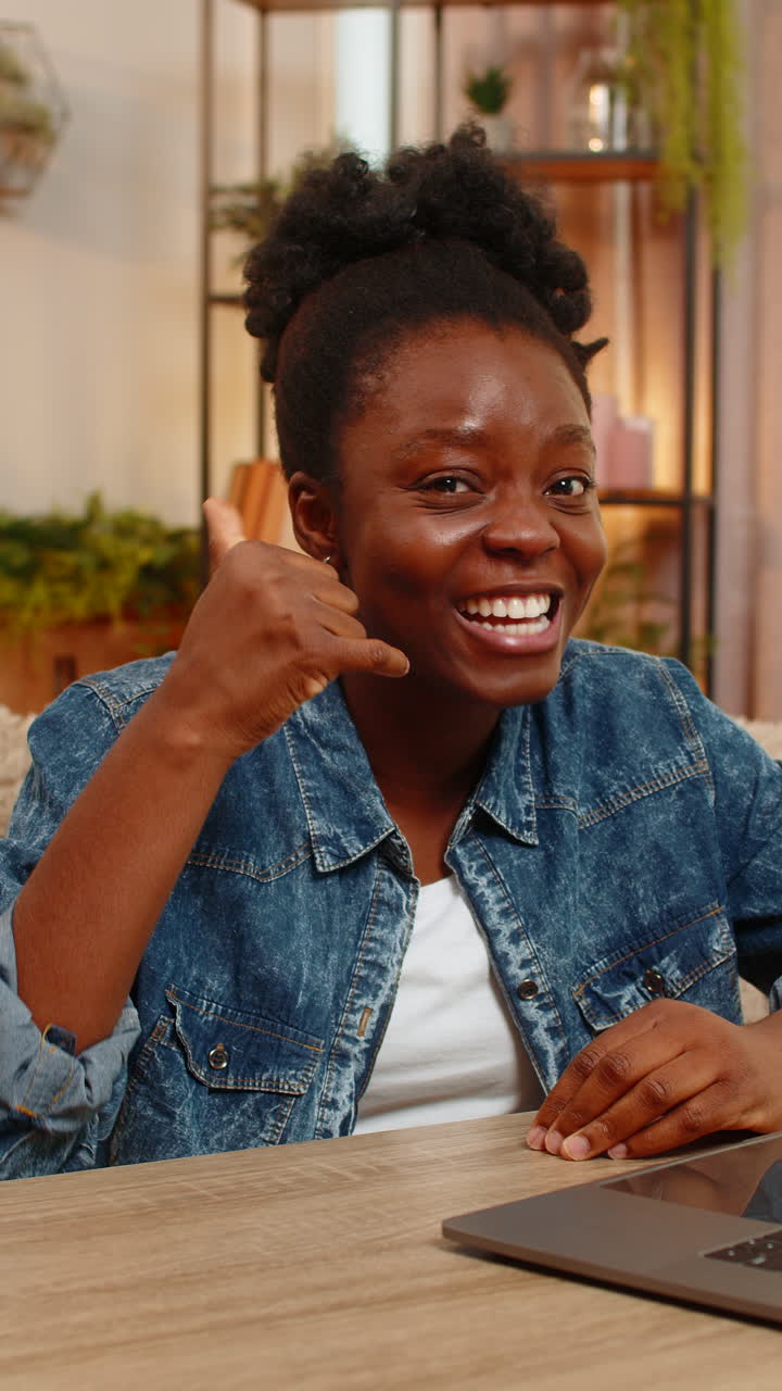 Giovane afroamericana civetta e mostra il gesto "chiamami" con la mano alla telecamera seduta a un tavolo a casa