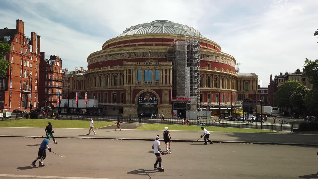 Royal Albert Hall with restoration scaffolding on facade and people playing roller hockey in the foreground, showing typical activity in London parks. Shot in Summer, Kensington Gardens, London.