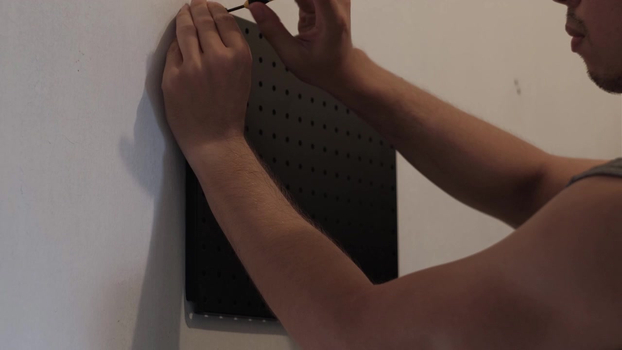 Medium static shot of a person mounting a black pegboard onto an interior wall using hand tools during home improvement work