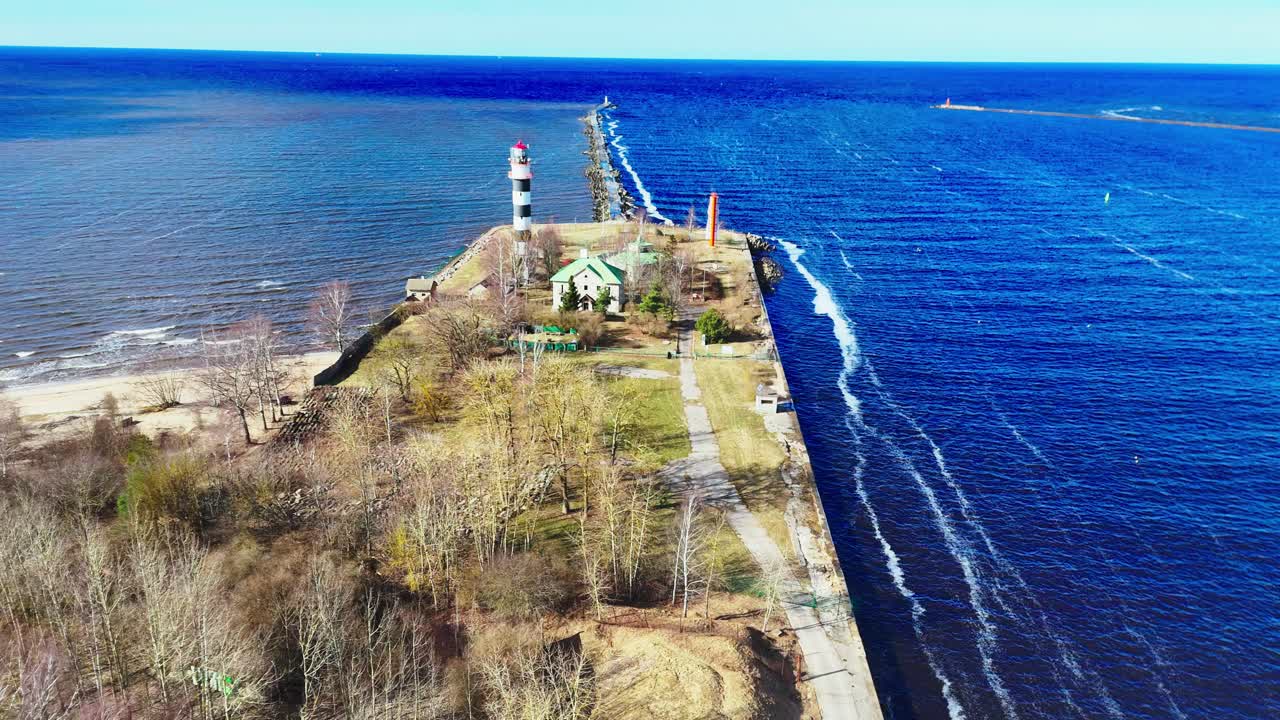 Aerial shot of Riga Lighthouse, Latvia, on a bright, clear spring day, showcasing the distinctive black and white tower long pier dividing deep blue and turquoise Baltic Sea waters