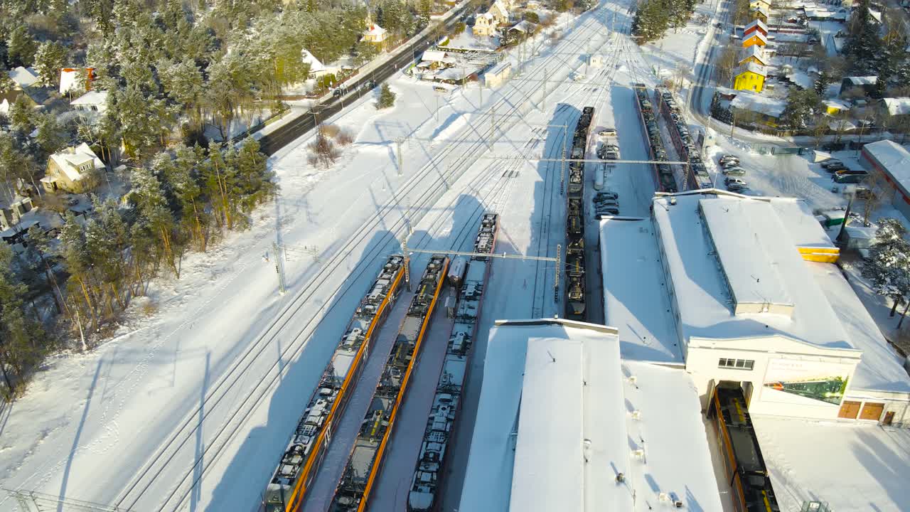 Aerial backward glide revealing a modern electric train depot with parked trains, snow covered warehouses, and long rail tracks stretching into the winter landscape under bright sunlight
