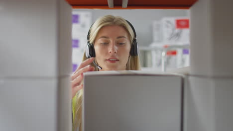 Female-Worker-Wearing-Headset-In-Logistics-Distribution-Warehouse-Putting-Box-On-Shelf