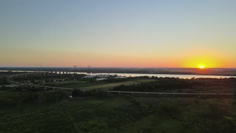 Dolly-right-flying-over-green-fields-and-Zarate-Brazo-Largo-road-and-railway-complex-cable-stayed-bridge-crossing-Parana-river-at-sunset,-Entre-Rios,-Argentina